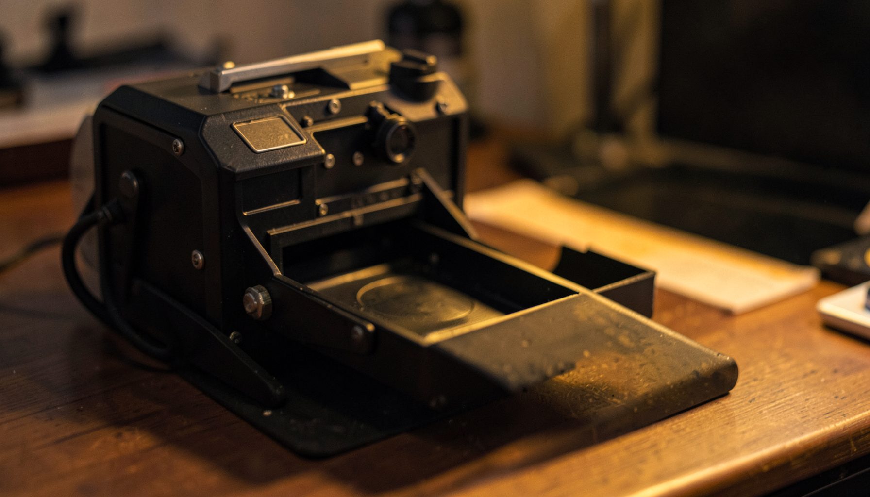 Close-up of a film scanner with a mounted slide tray on a desk, warm amber and charcoal tones, precise technical mood