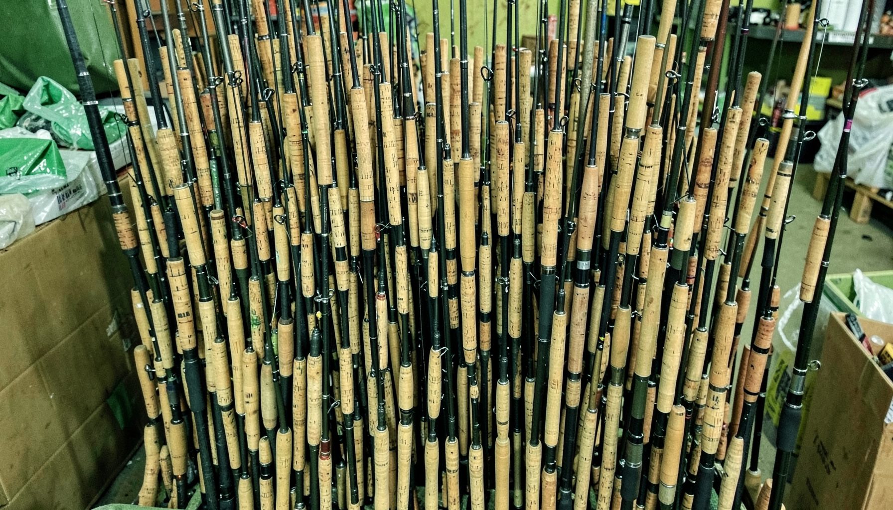Dense cluster of fishing rods with cork handles standing upright in storage, green and tan hues