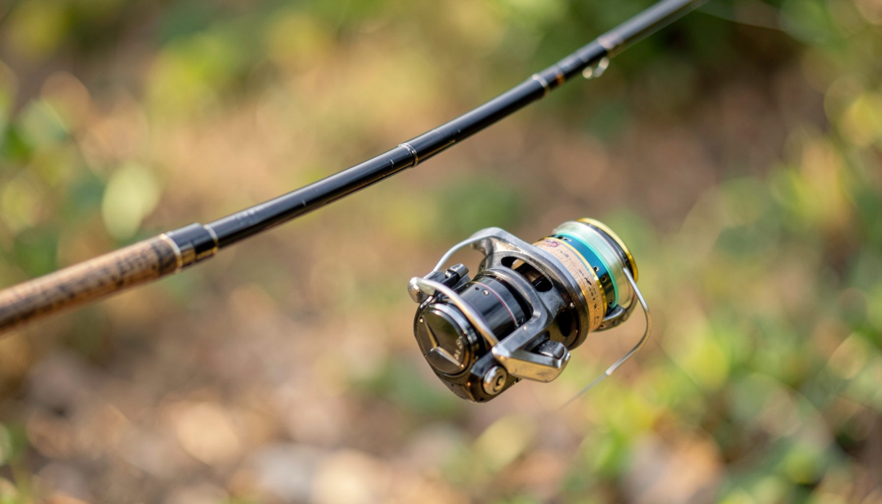 Close-up of a fishing reel attached to a rod tip against a blurred green and brown natural background, warm outdoor mood